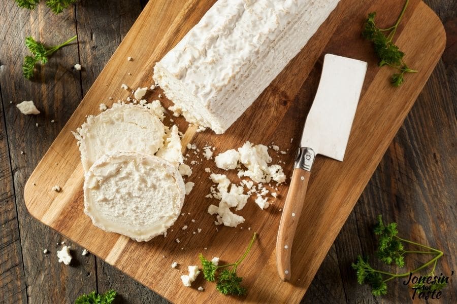 a log of goat cheese on a cutting board.  A couple slices sit next to a cleaver style knife and some crumbled goat cheese.