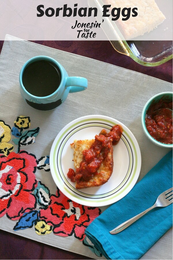 A slice of Sorbian Eggs topped with salsa on white plate on a bold flower placemat with a blue mug.