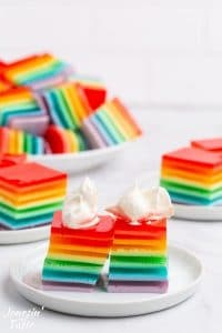 two squares of rainbow jello on a white plate each topped with a dollop of whipped cream
