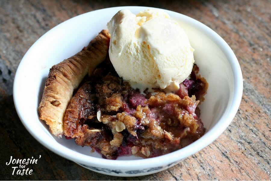 A slice of apple crumble pie topped with a scoop of vanilla ice cream in a white bowl