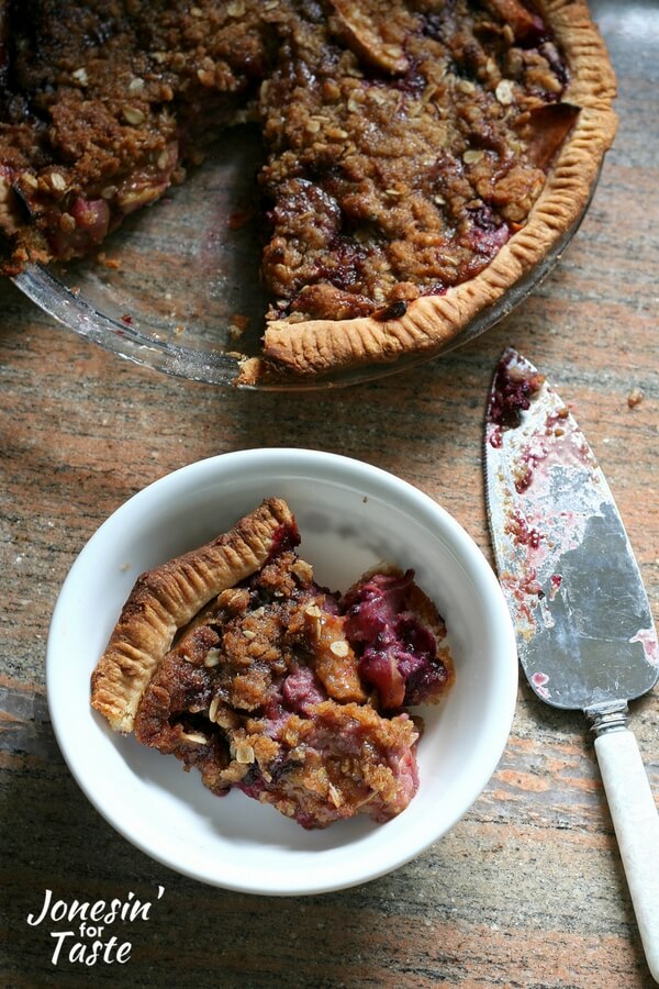 A slice of apple berry pie in a bowl sitting next to the whole pie with a pie server next to both.