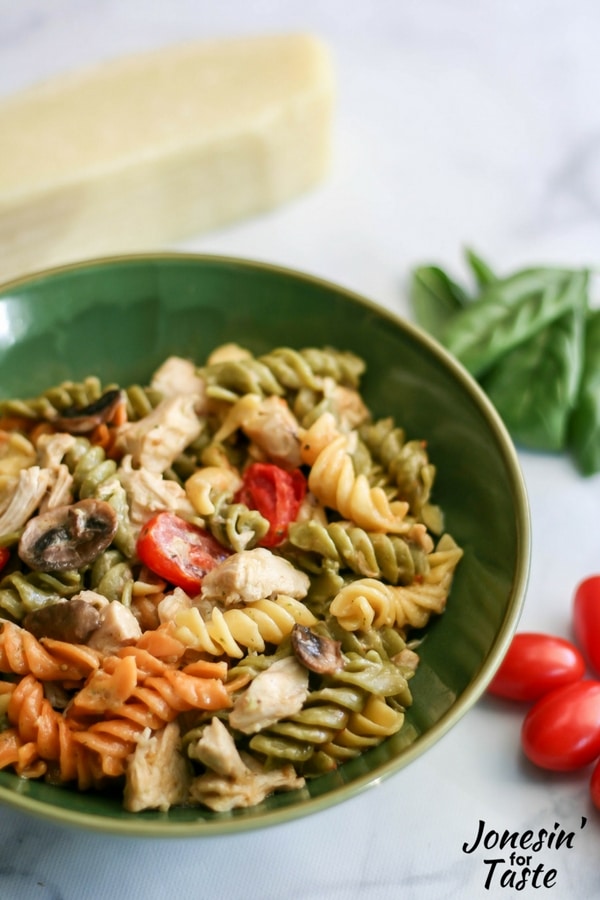 A green bowl filled with pesto chicken alfredo surrounded by groups of basil leaves, cherry tomatoes, and a hunk of parmesan cheese.
