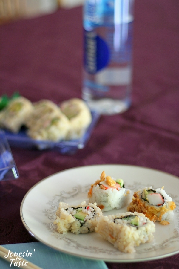 A plate of sushi on a purple tablecloth with a bottle of water and a container of sushi in the background