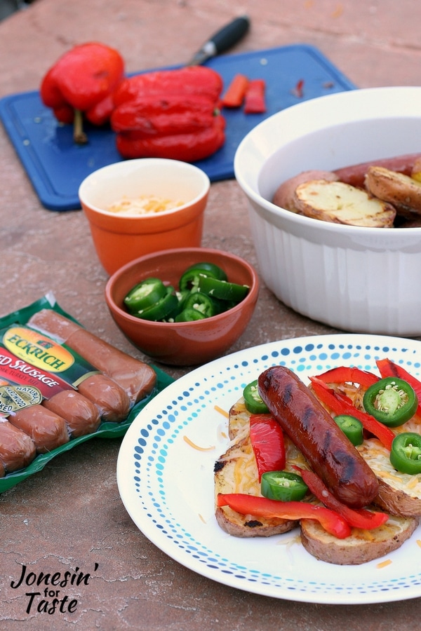 A plate of grilled smoked sausage and potatoes with toppings with a package of smoked sausages, and bowls of toppings surrounding the plate with a dish of grilled potatoes in the background