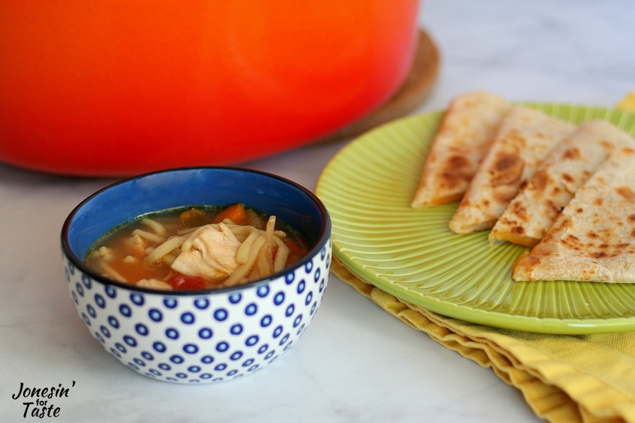 A blue bowl of quick salsa soup with an orange pot and green plate with quesadillas in the background