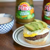 A Huevos Rancheros Burger topped with avocado served up with a dish a salsa spread with 2 bottles of Pace Salsa in the background.