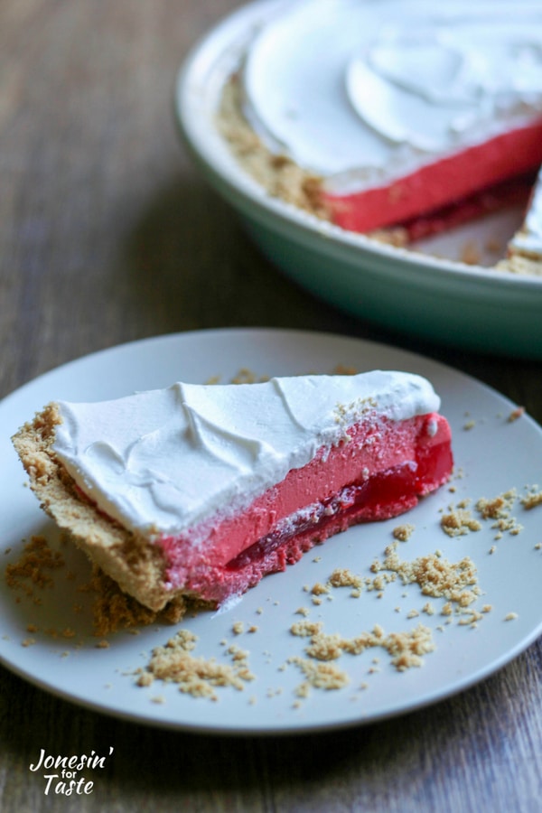 A slice of easy raspberry jello pie with a graham cracker crust on a plate in front of a pie plate with the rest of the pie.