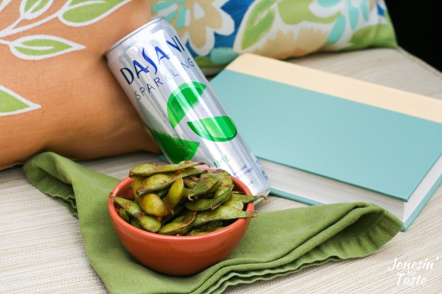 A bowl of garlic soy edamame next to a can of DASANI sparkling next to a book on a chair