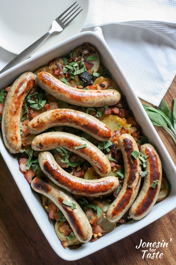 a white casserole dish filled with sliced potatoes, bacon, herbs, and browned bratwurst sausages
