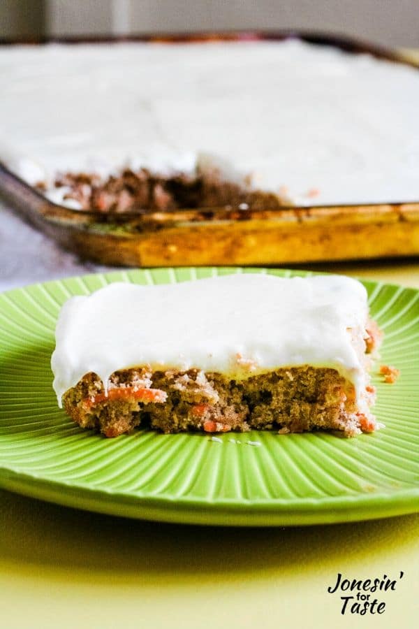 a slice of carrot sheet cake covered with cream cheese icing on a plate next to a cookie sheet