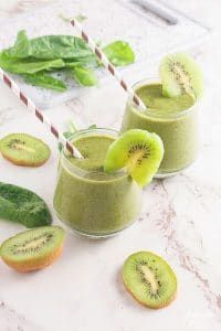 2 cups of kiwi smoothie with a slice of kiwi and a straw in each surrounded by halved kiwis and spinach leaves