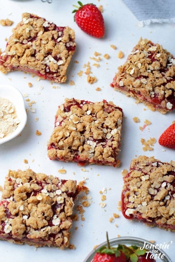 5 squares of strawberry bars sitting on a white background with crumbs and fresh strawberries artfully arranged around them