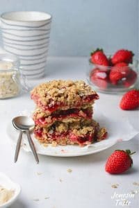 3 strawberry oatmeal crumble bars stacked on a white plate with containers of fresh strawberries and oatmeal around it