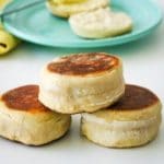 one English muffin is stacked on top of another 2 on a white table. A cut muffin is in the background on a blue plate